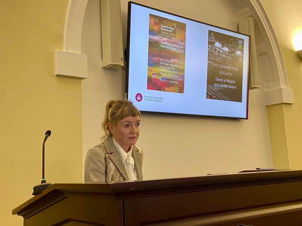 A woman presents from a podium. Text in Irish language is on screen behind her