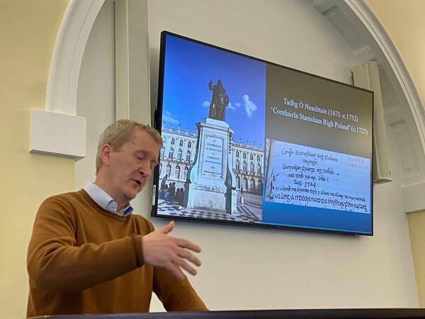 A man presents from a podium. Text in Irish language is on screen behind him